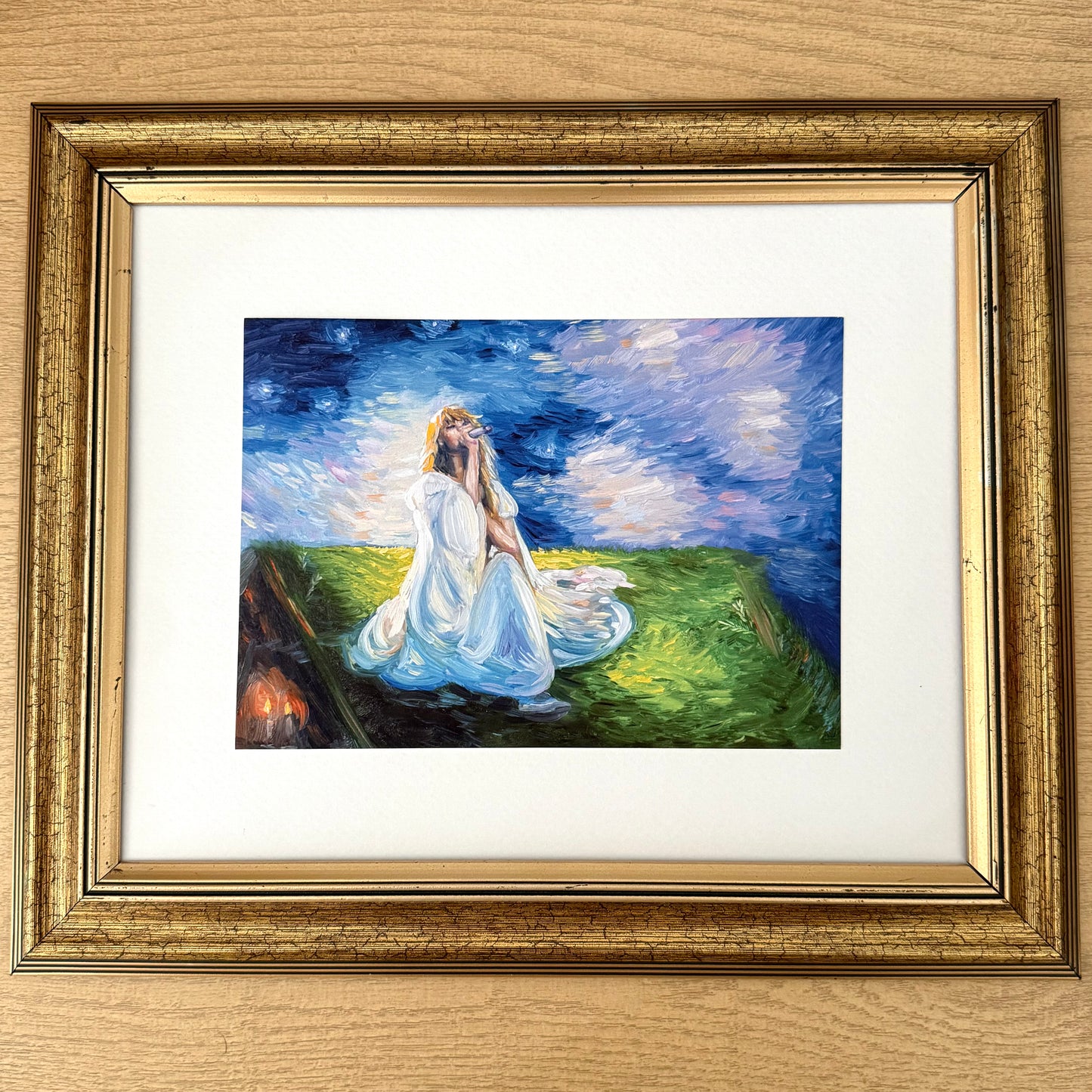 A framed impressionist painting of a woman in a white dress kneeling on a grassy hill under a starry night sky, presented in a distressed gold frame with a white mat on a light wood background.