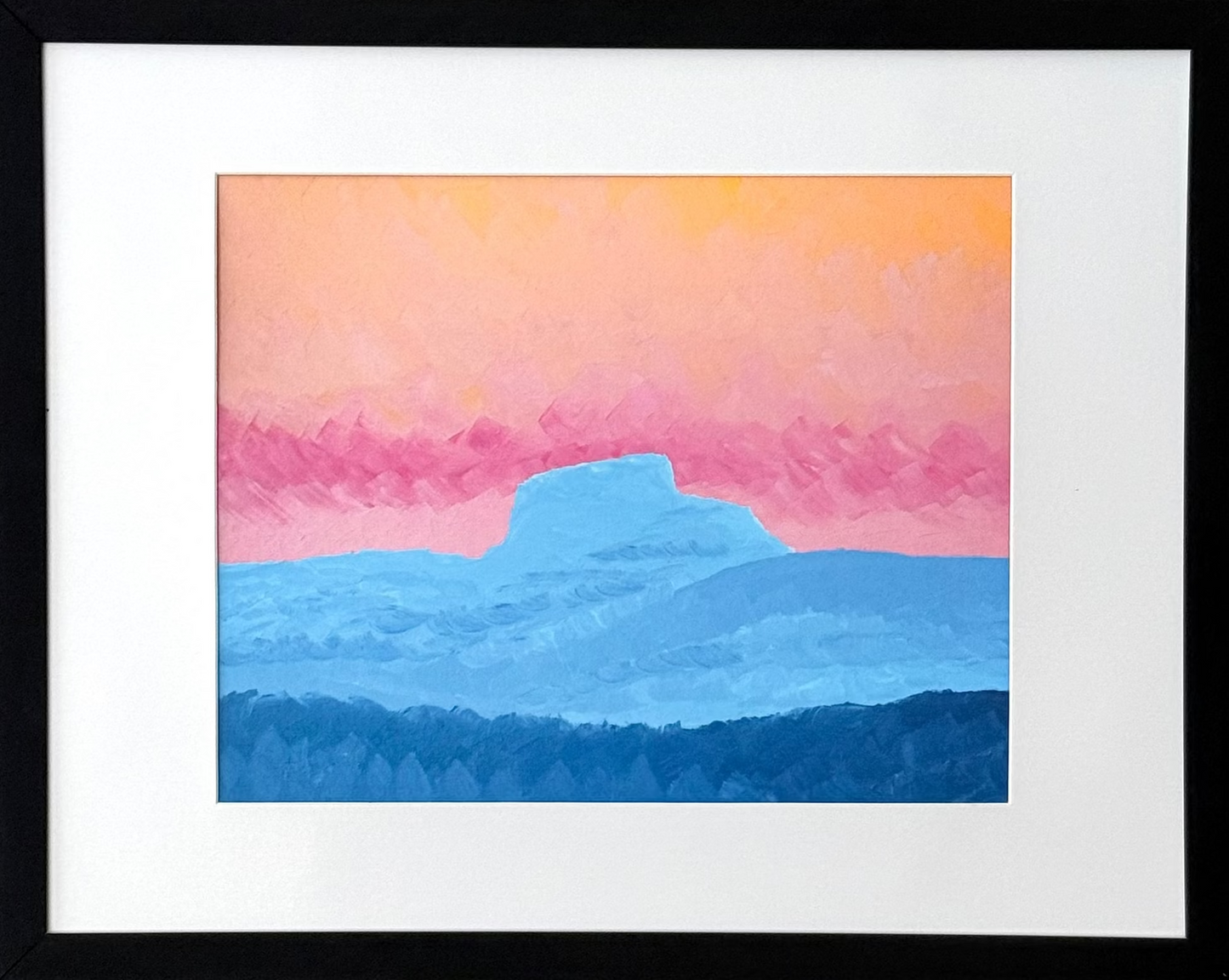 A framed landscape art print featuring a stylized sunset or sunrise with a pink and orange sky, blue mountains, and a dark blue foreground, displayed on a light gray wall.