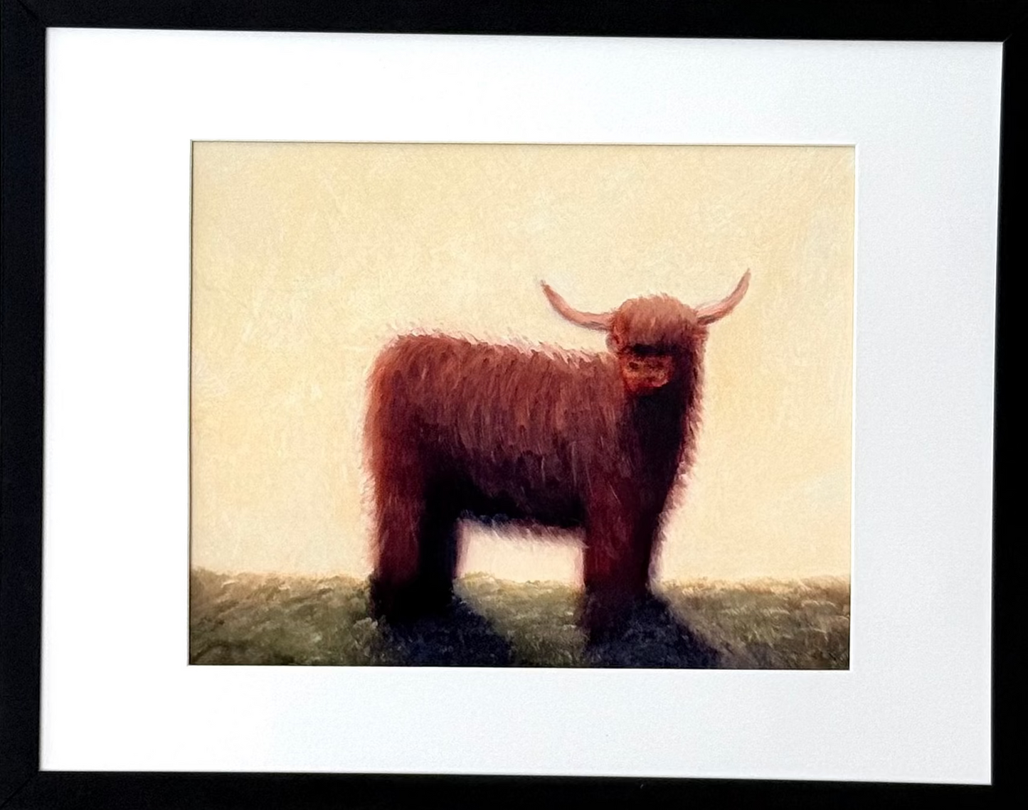 A framed art print featuring a brown Highland cow illustration on a cream background, set in a black frame with a white mat against a light gray wall.