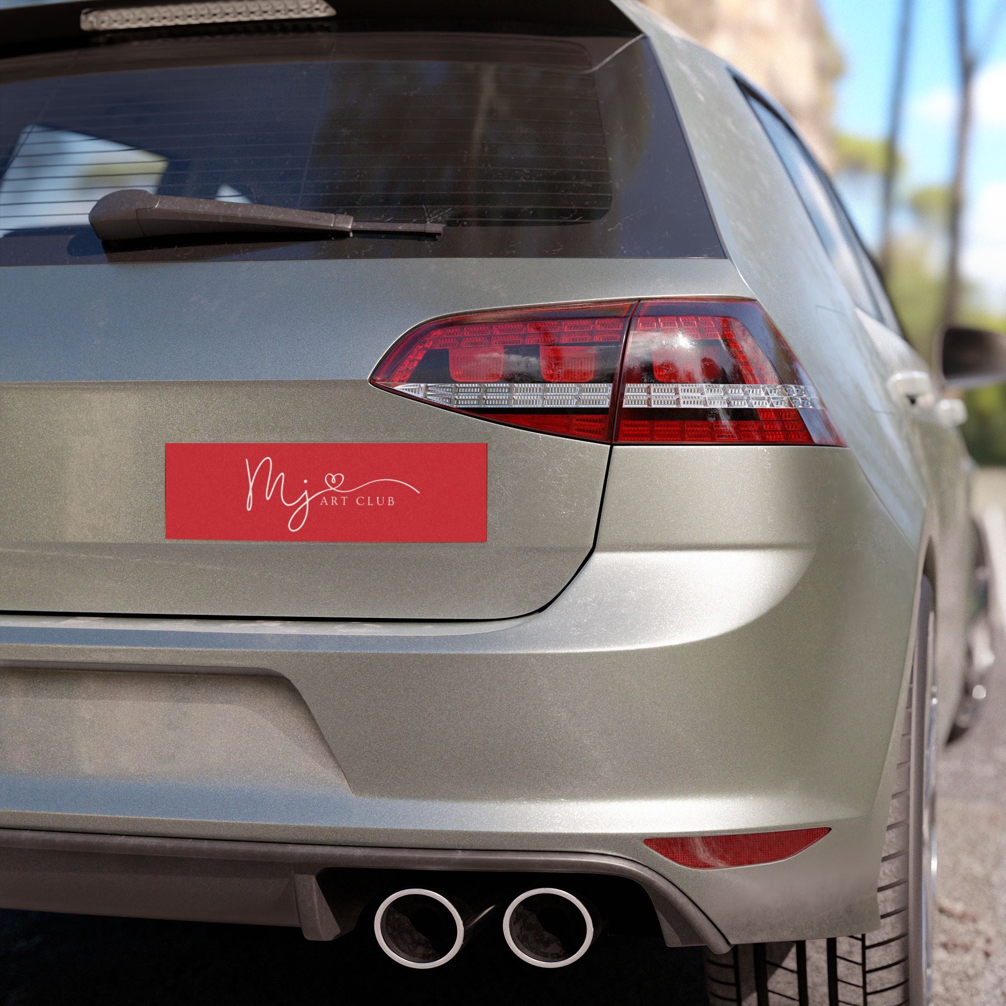 A red rectangular vinyl decal sticker with white script text reading 'MJ Art Club' applied to the rear left side of a silver hatchback car, shown in an outdoor setting.