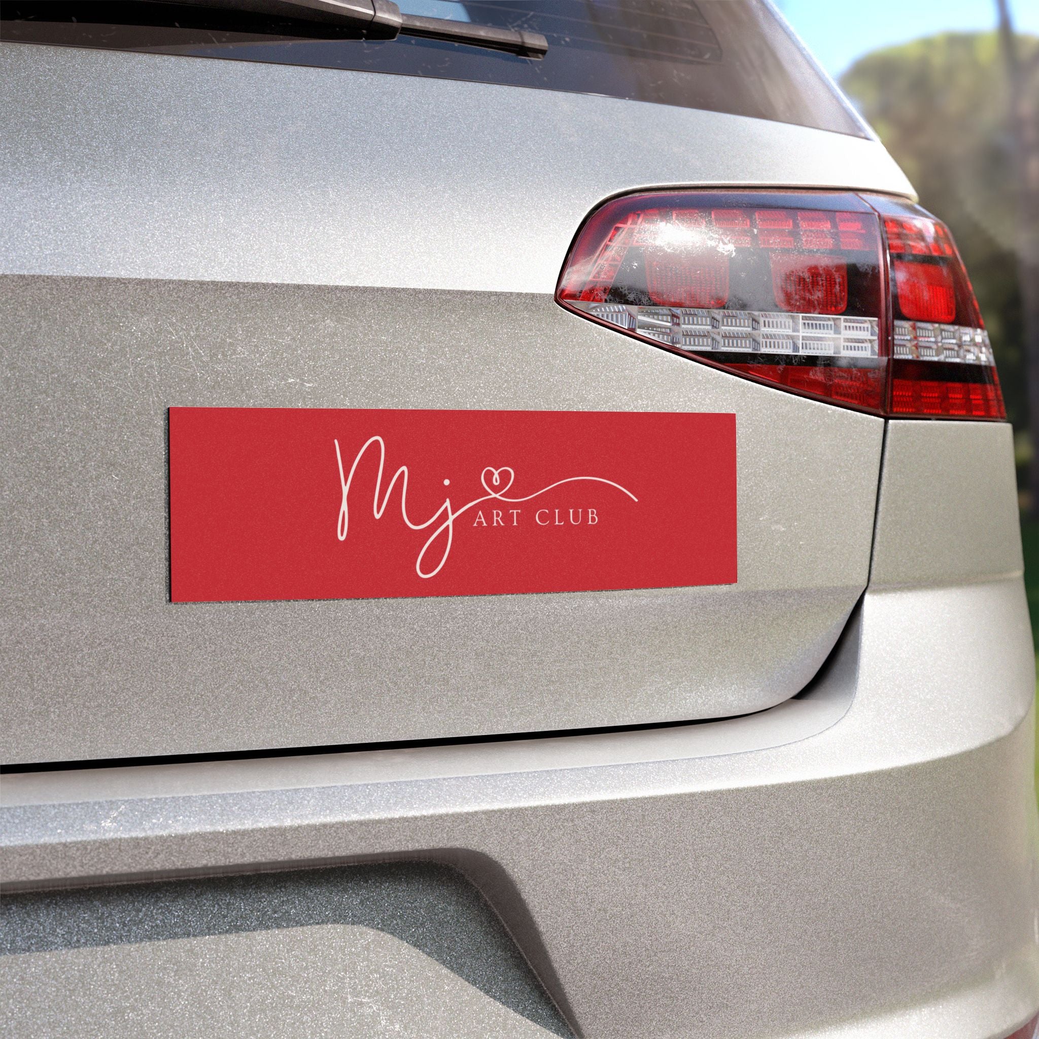 A red rectangular car window decal with white script reading 'Mj ART CLUB' and a small heart, shown applied to the rear of a silver vehicle.