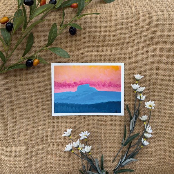 Burlap background with various floral decorations surrounding a fine art print depicting blue mountains with a yellow and pink sunset