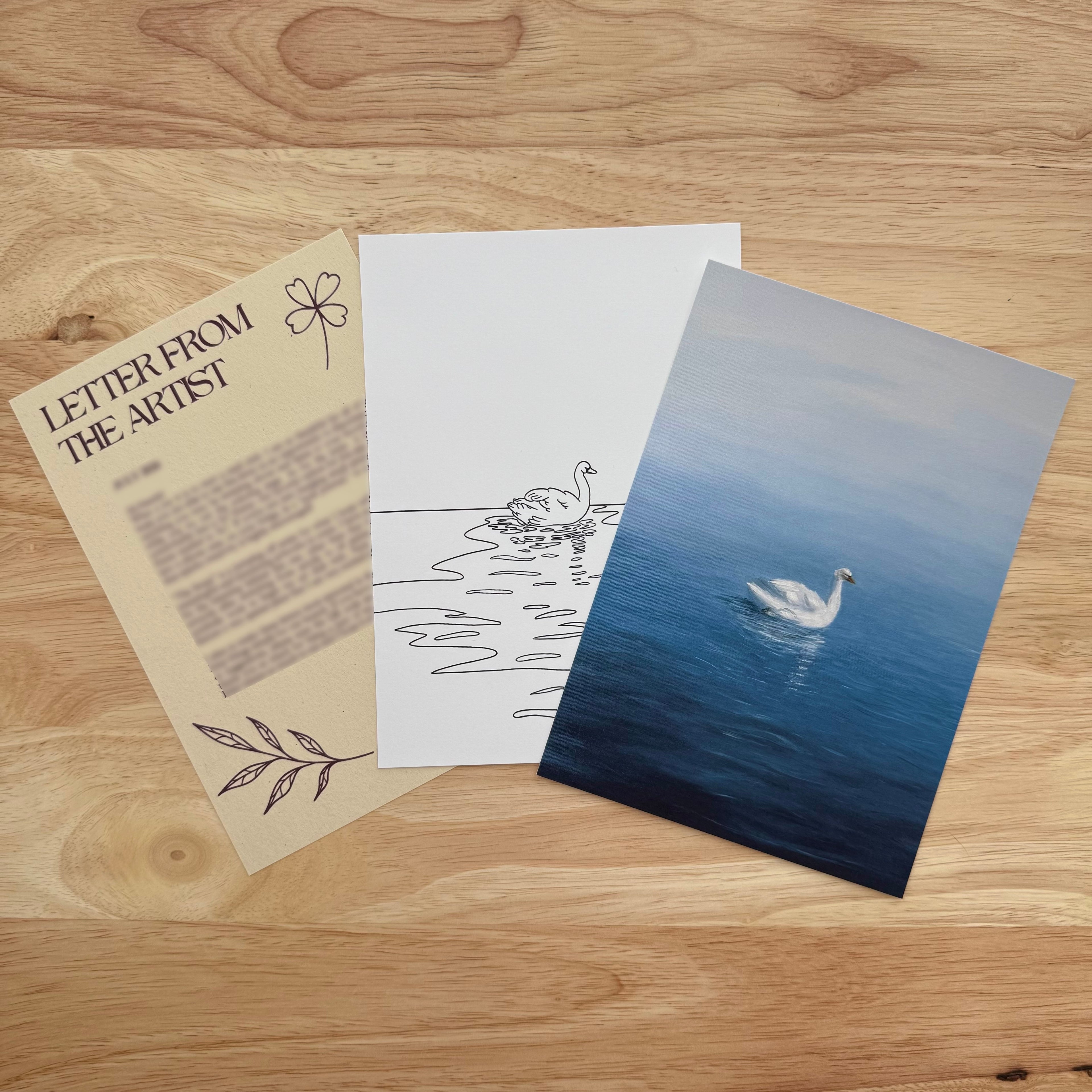 Three art prints or cards arranged on a light wood tabletop: a beige card reading 'LETTER FROM THE ARTIST' with text and line drawings, a white card with a minimalist swan sketch, and a blue-toned photo print of a swan on water.