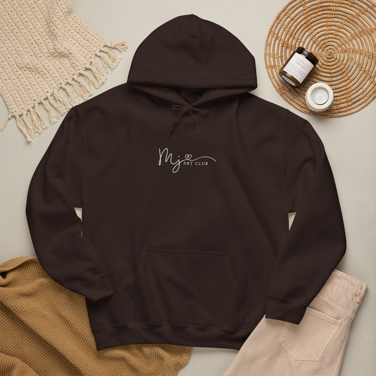 Brown hoodie with a logo on a light gray surface with beige pants and a woven mat.
