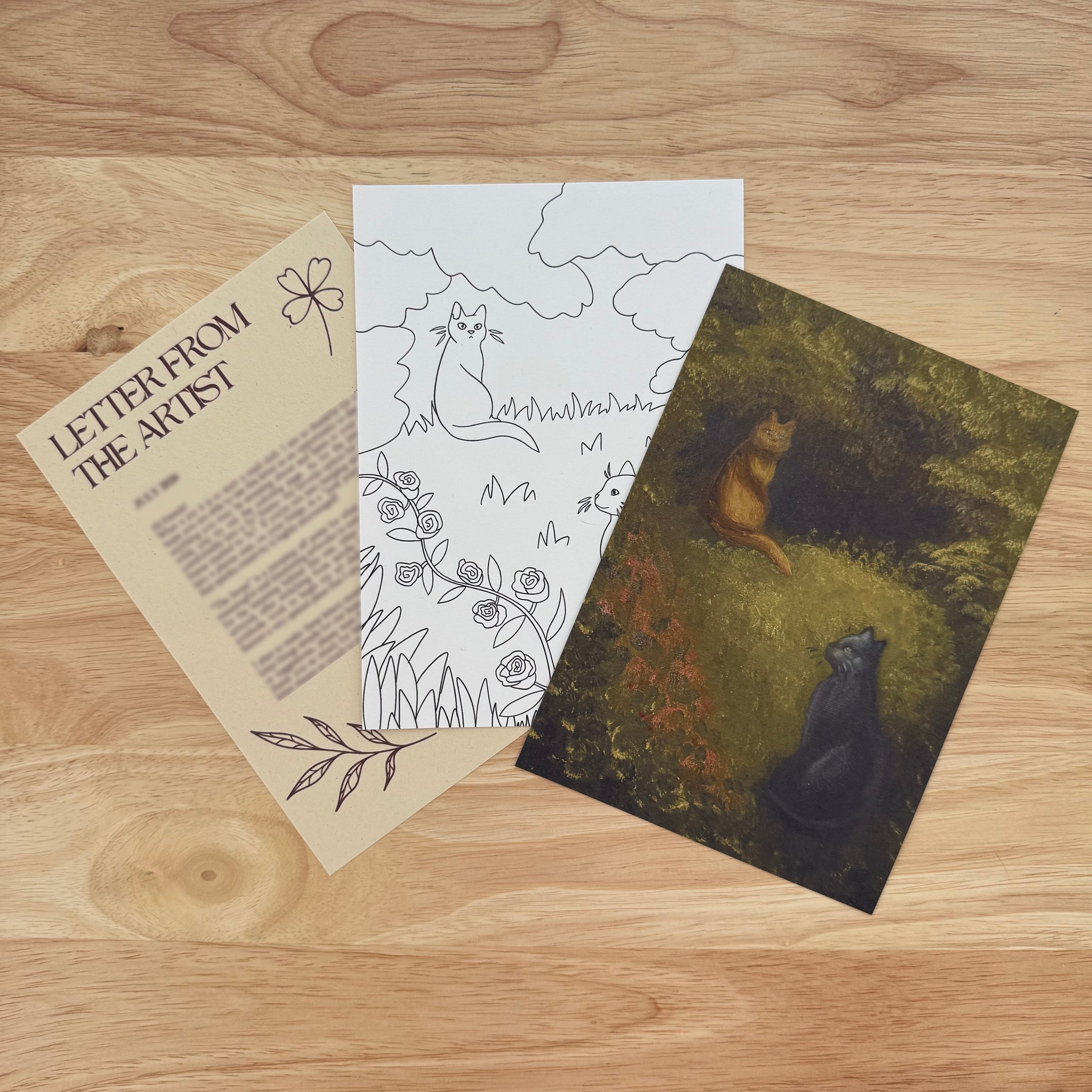 Three art prints fanned out on a light wood tabletop. The left print is beige with the text 'LETTER FROM THE ARTIST' and line drawings. The middle print is a coloring page featuring two cats. The final print is a painting of two cats.