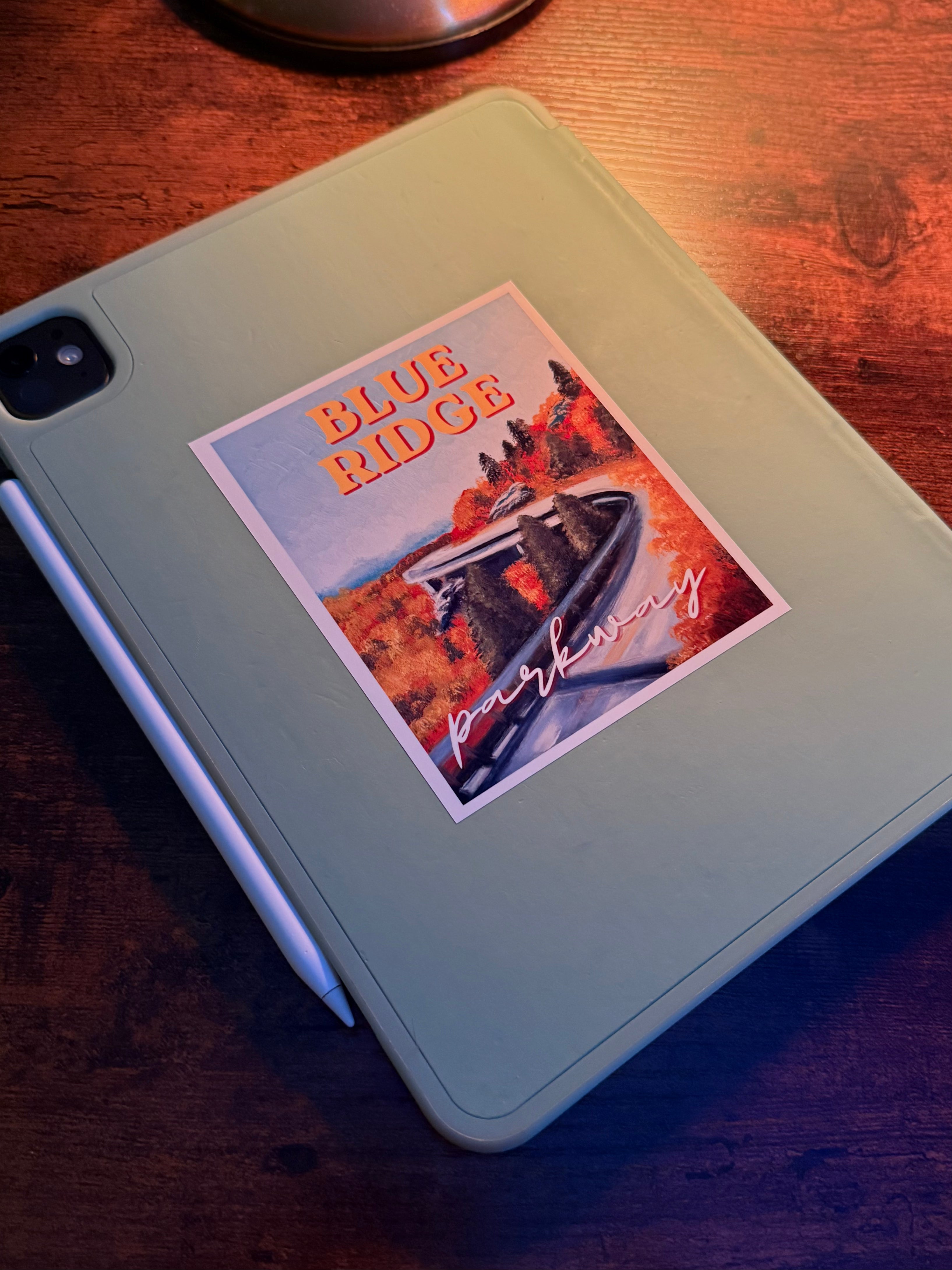 A sage green tablet case with a square sticker that reads 'BLUE RIDGE parkway' in orange and white, resting on a dark wooden table with a white stylus.