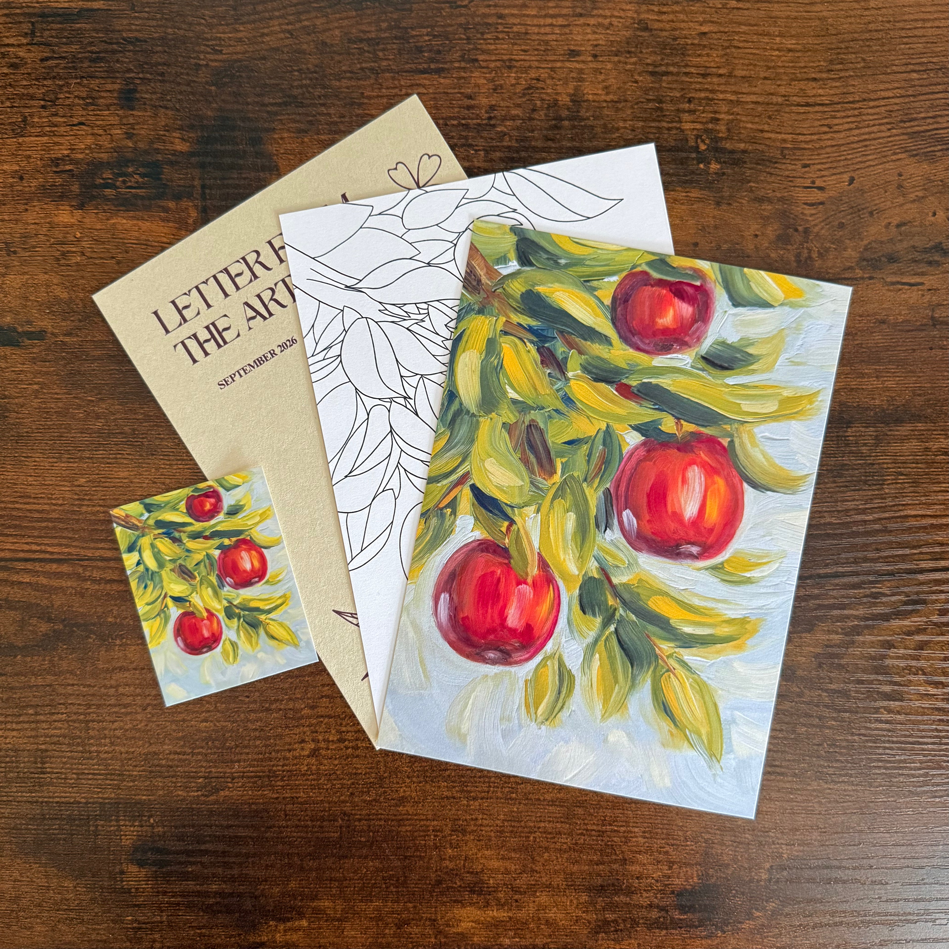 An autumn-themed art letter set featuring a main letter card with a painted red apple branch, a matching coloring page and small rectangle sticker, and a brown letter with 'LETTER FROM THE ARTIST SEPTEMBER 2026' printed on it, arranged on a dark wood background.