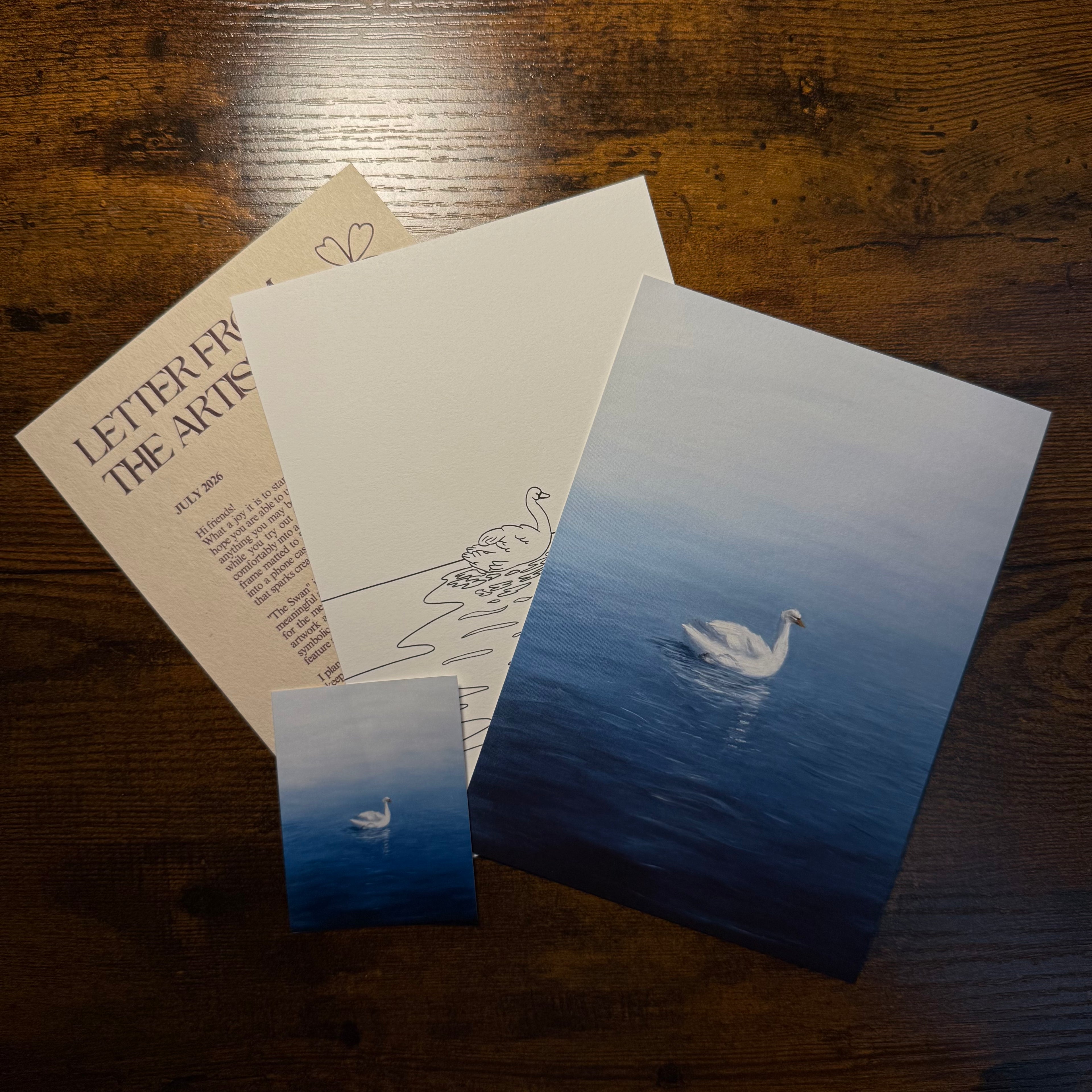 A set of swan-themed art prints and stationery laid out on a dark wooden table. The set includes a large square print, a smaller rectangular print, a blank card with a swan line drawing, and a beige card partially showing the text 'LETTER FROM THE ARTIST'.