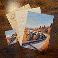 A set of art prints and postcards featuring a painted autumn landscape with a winding road, trees, mountains, and a bridge, displayed on a dark wooden table.