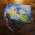 A stack of art prints or postcards featuring an impressionistic painting of a woman in a flowing white dress sitting on a grassy hill under a starry blue sky, displayed on a dark wooden tabletop.