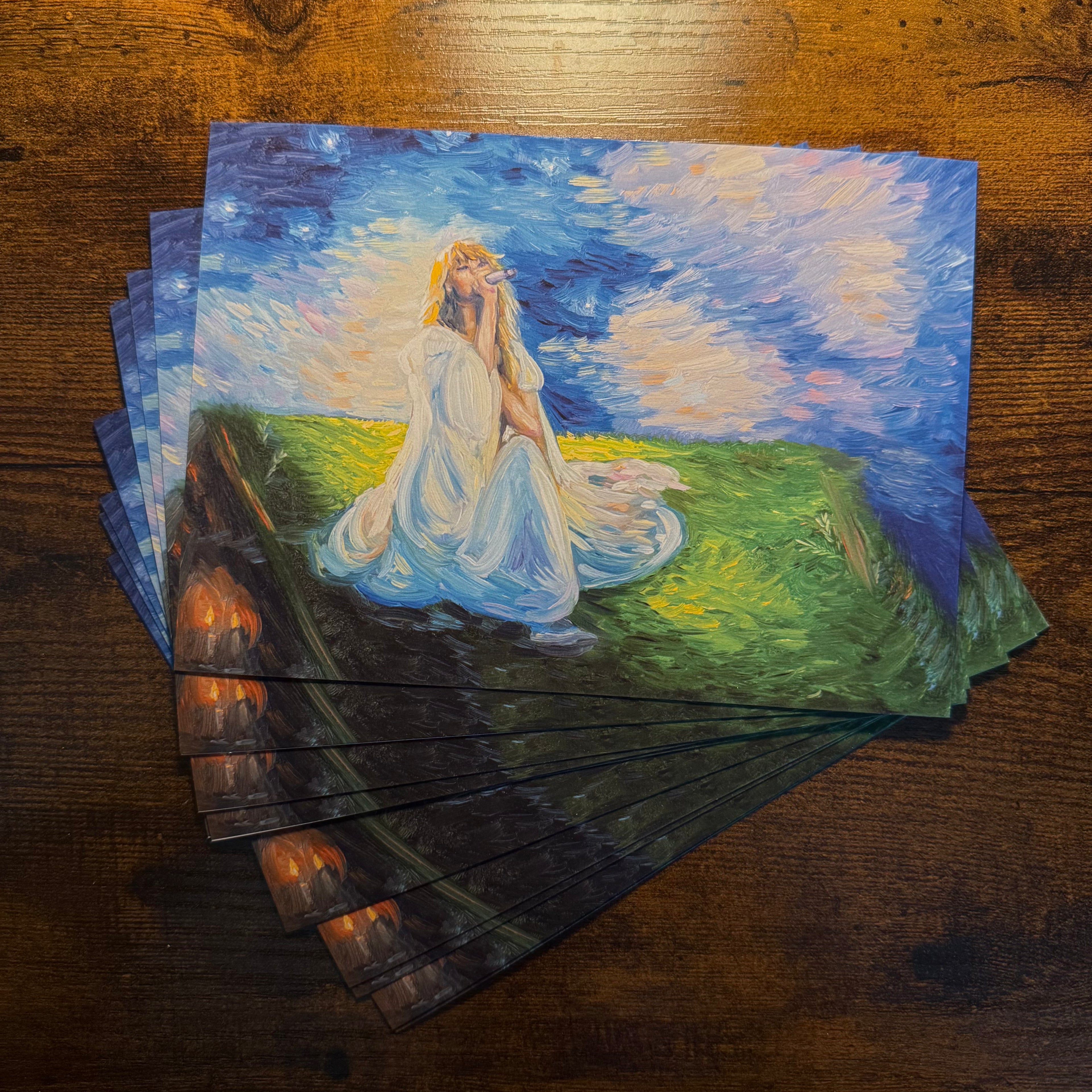 A stack of art prints or postcards featuring an impressionistic painting of a woman in a flowing white dress sitting on a grassy hill under a starry blue sky, displayed on a dark wooden tabletop.