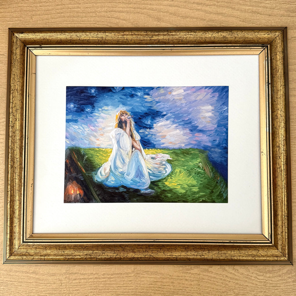 A framed impressionist painting of a woman in a white dress kneeling on a grassy hill under a starry night sky, presented in a distressed gold frame with a white mat on a light wood background.