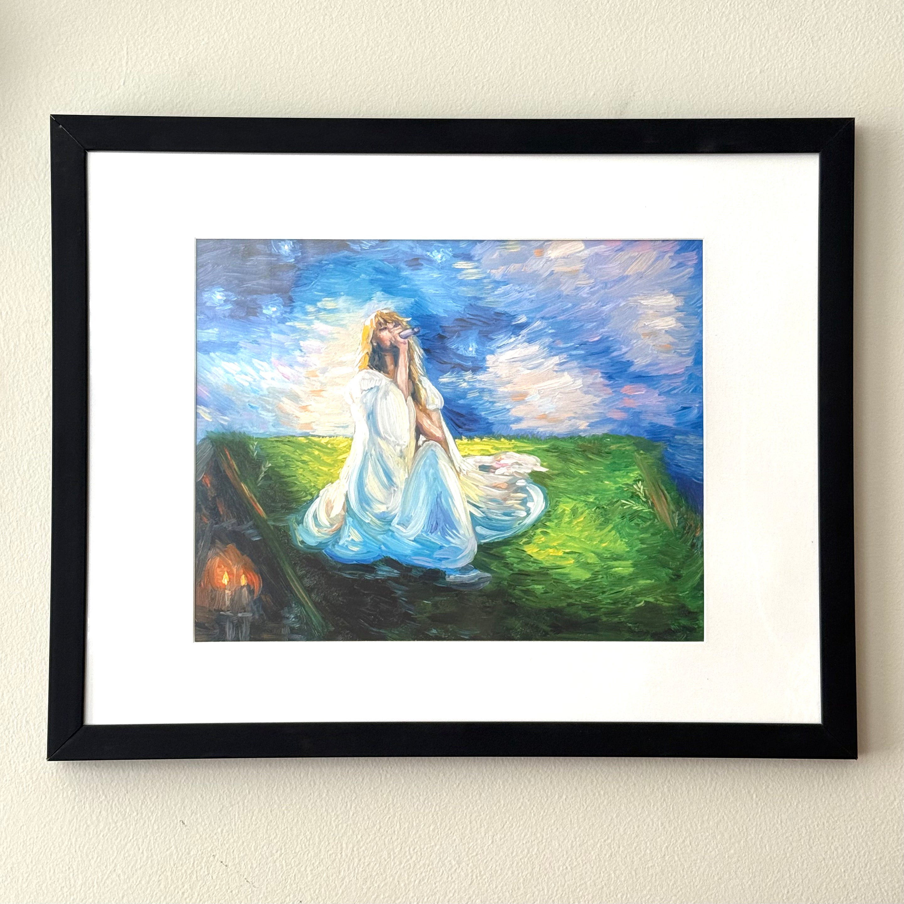 A framed art print featuring an impressionistic painting of a woman in a flowing white dress sitting on a green hill under a blue sky, mounted on a textured beige wall.