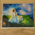 An art print of a painting titled 'TAKE HER HOME' displayed on a light wood floor. The painting depicts a woman in a flowing white gown sitting in a grassy field under a starry night sky, with a small set of candles burning in the background.