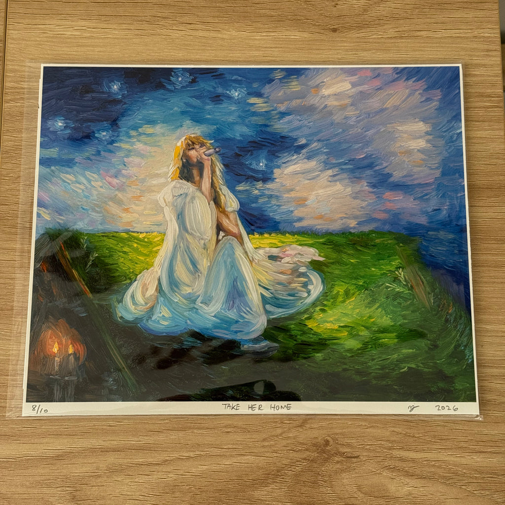 An art print of a painting titled 'TAKE HER HOME' displayed on a light wood floor. The painting depicts a woman in a flowing white gown sitting in a grassy field under a starry night sky, with a small set of candles burning in the background.