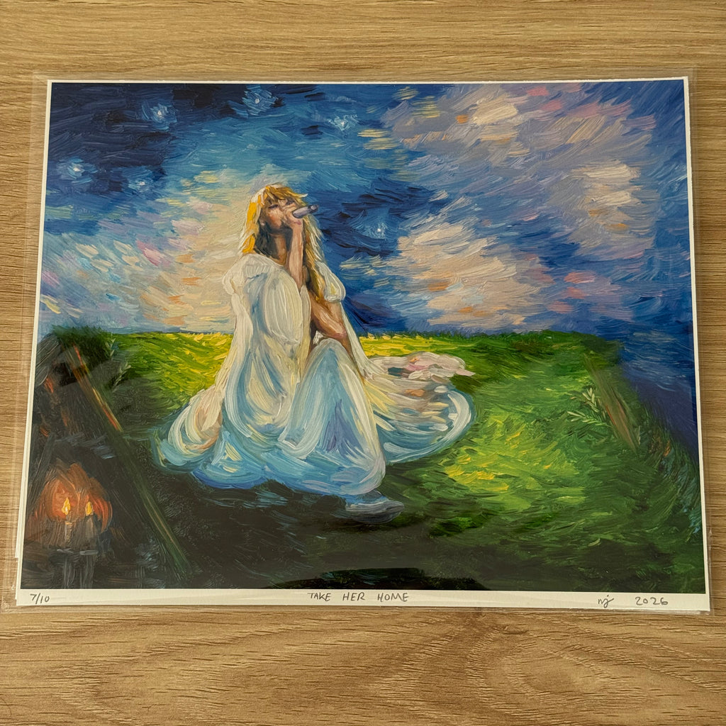 An art print of a painting titled 'TAKE HER HOME' displayed on a light wood floor. The painting depicts a woman in a flowing white gown sitting in a grassy field under a starry night sky, with a small set of candles burning in the background.