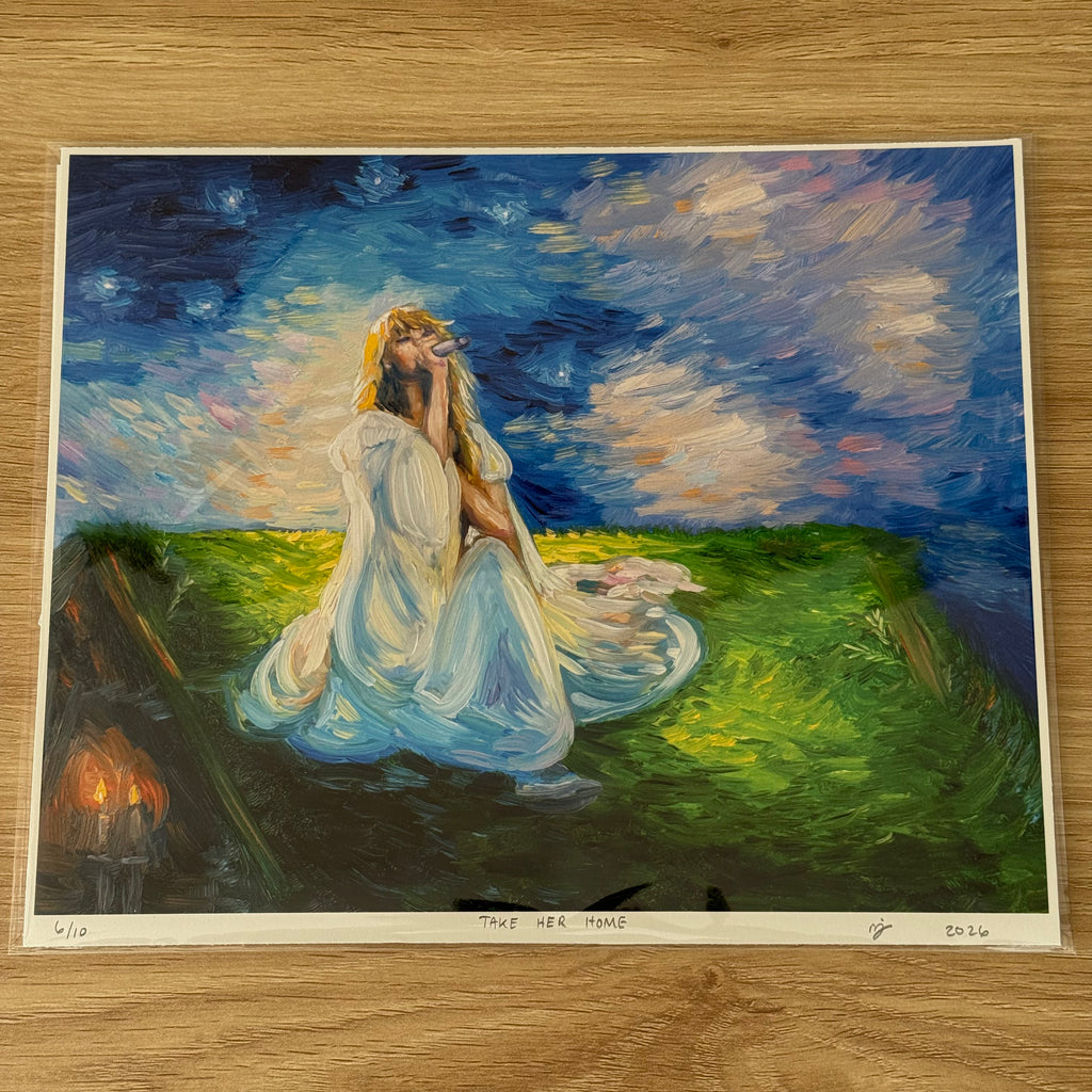 An art print of a painting titled 'TAKE HER HOME' displayed on a light wood floor. The painting depicts a woman in a flowing white gown sitting in a grassy field under a starry night sky, with a small set of candles burning in the background.