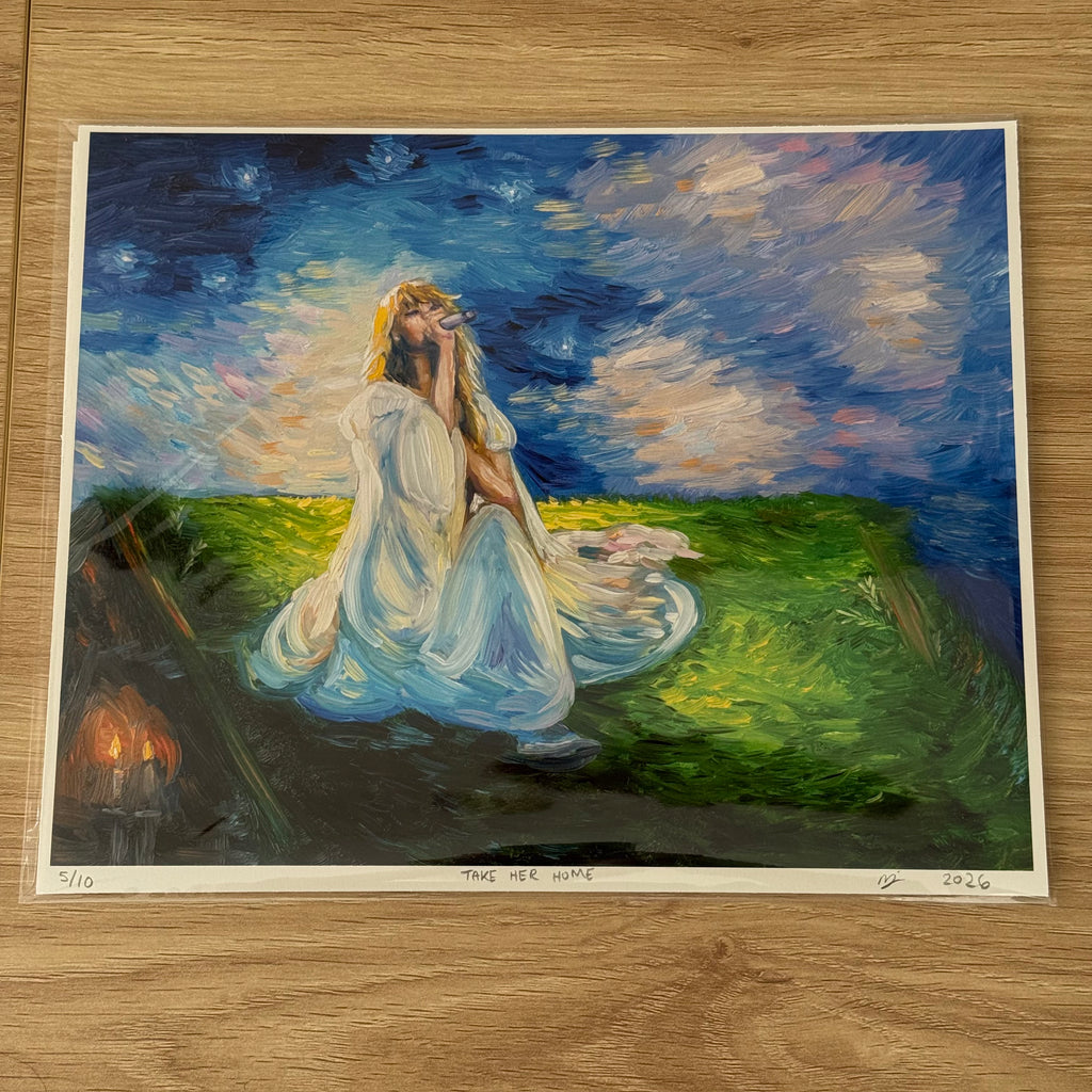 An art print of a painting titled 'TAKE HER HOME' displayed on a light wood floor. The painting depicts a woman in a flowing white gown sitting in a grassy field under a starry night sky, with a small set of candles burning in the background.