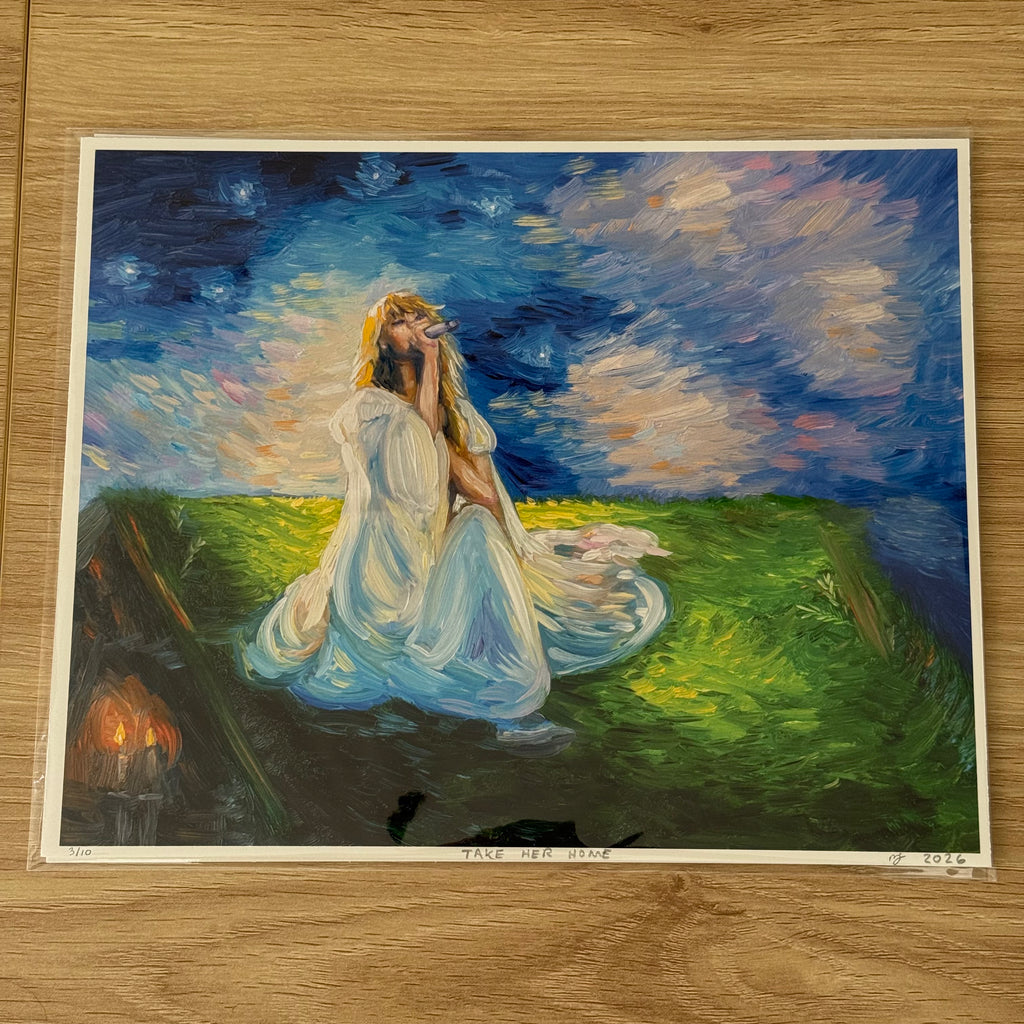 An art print of a painting titled 'TAKE HER HOME' displayed on a light wood floor. The painting depicts a woman in a flowing white gown sitting in a grassy field under a starry night sky, with a small set of candles burning in the background.