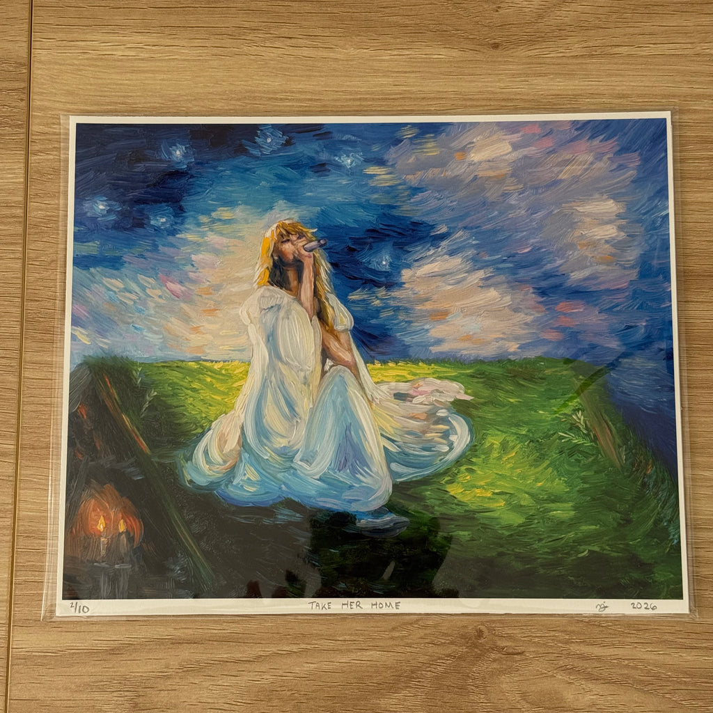 An art print of a painting titled 'TAKE HER HOME' displayed on a light wood floor. The painting depicts a woman in a flowing white gown sitting in a grassy field under a starry night sky, with a small set of candles burning in the background.