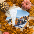 A flat lay of three art cards and a sticker, all fall mountain road themed, surrounded by orange leaves and faux pumpkins on a wood background.