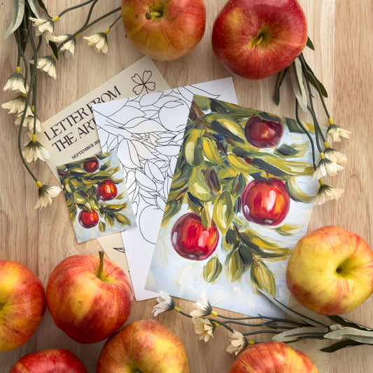 Photo of a letter, a coloring page, a sticker, and a fine art print, all apple-themed, on a wood background surrounded by apples and flowers.