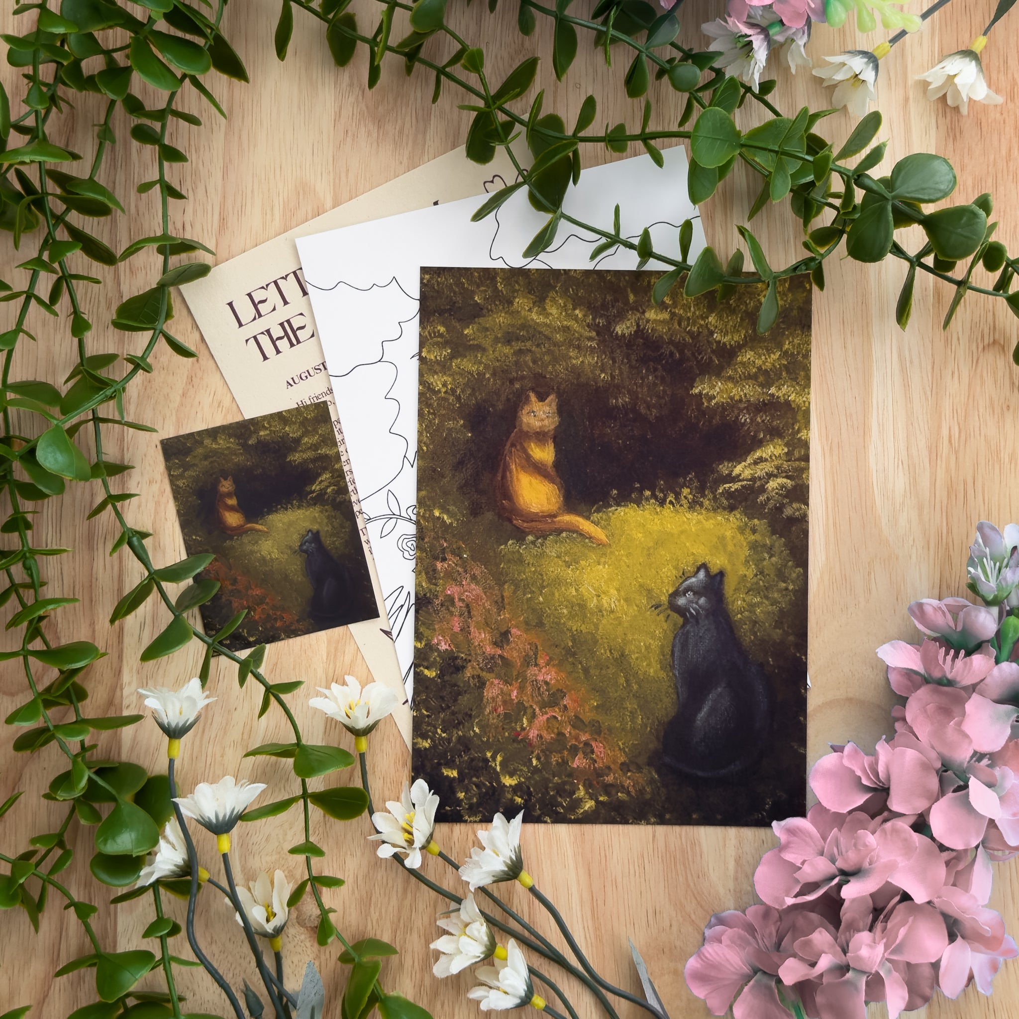 A flat-lay photograph of art prints featuring a forest scene with two cats, styled with green foliage and pink flowers on a light wood background. The main print shows a black cat and an orange tabby cat in a forest clearing.