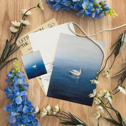 A flat lay with three swan-themed art cards and a sticker on a wood background with flowers and a pearl necklace