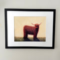 Framed art print featuring a painted Highland cow standing on a grassy field against a pale cream background, matted with white and enclosed in a black frame, mounted on a light wall.