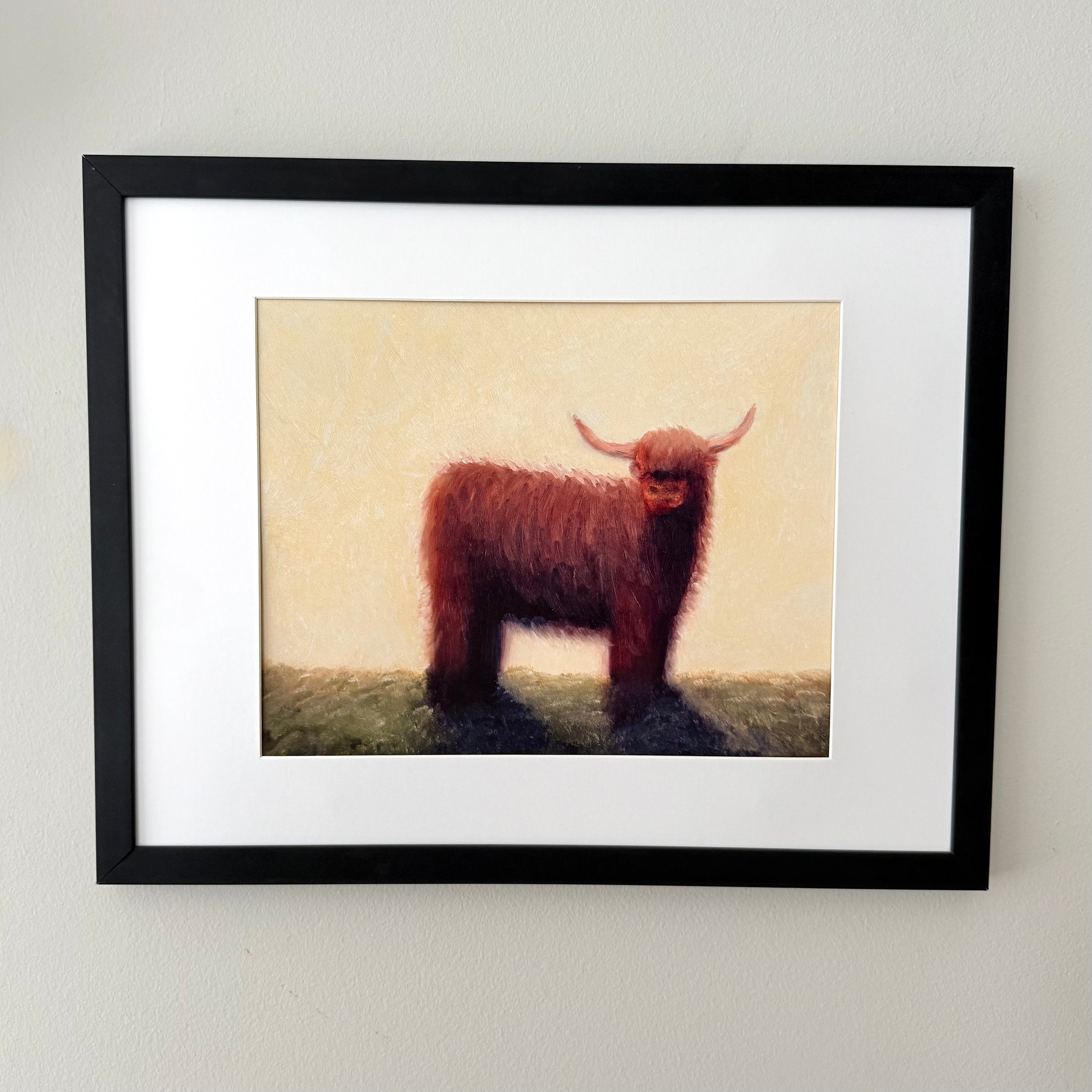 Framed art print featuring a painted Highland cow standing on a grassy field against a pale cream background, matted with white and enclosed in a black frame, mounted on a light wall.