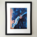 Framed wall art featuring an expressive blue painting of a person playing an electric guitar, shown in a black frame with a white mat against a light beige wall.