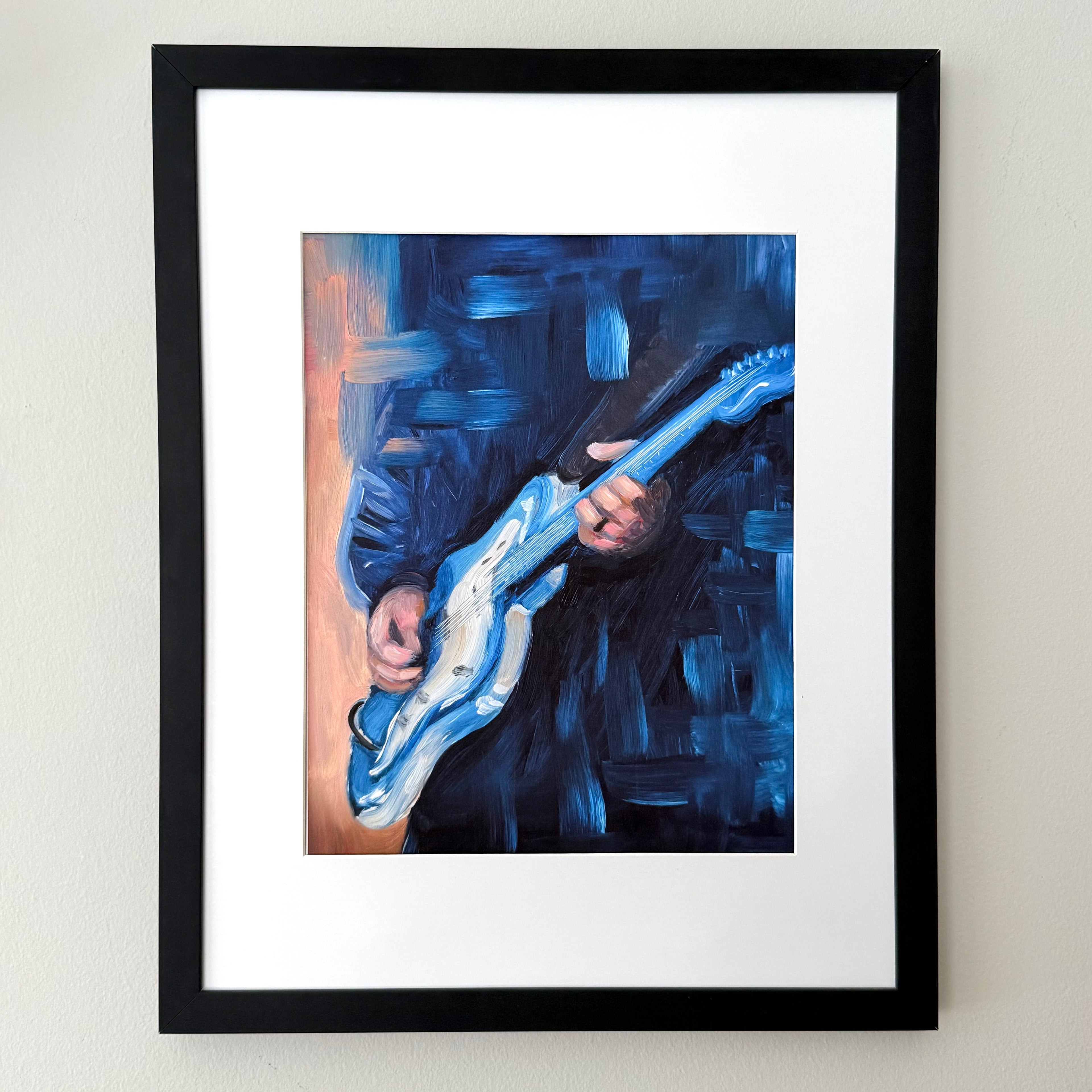 Framed wall art featuring an expressive blue painting of a person playing an electric guitar, shown in a black frame with a white mat against a light beige wall.