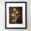 Framed floral wall art featuring three yellow tulips on a dark brown background, mounted with a white mat and a black frame on a light wall.