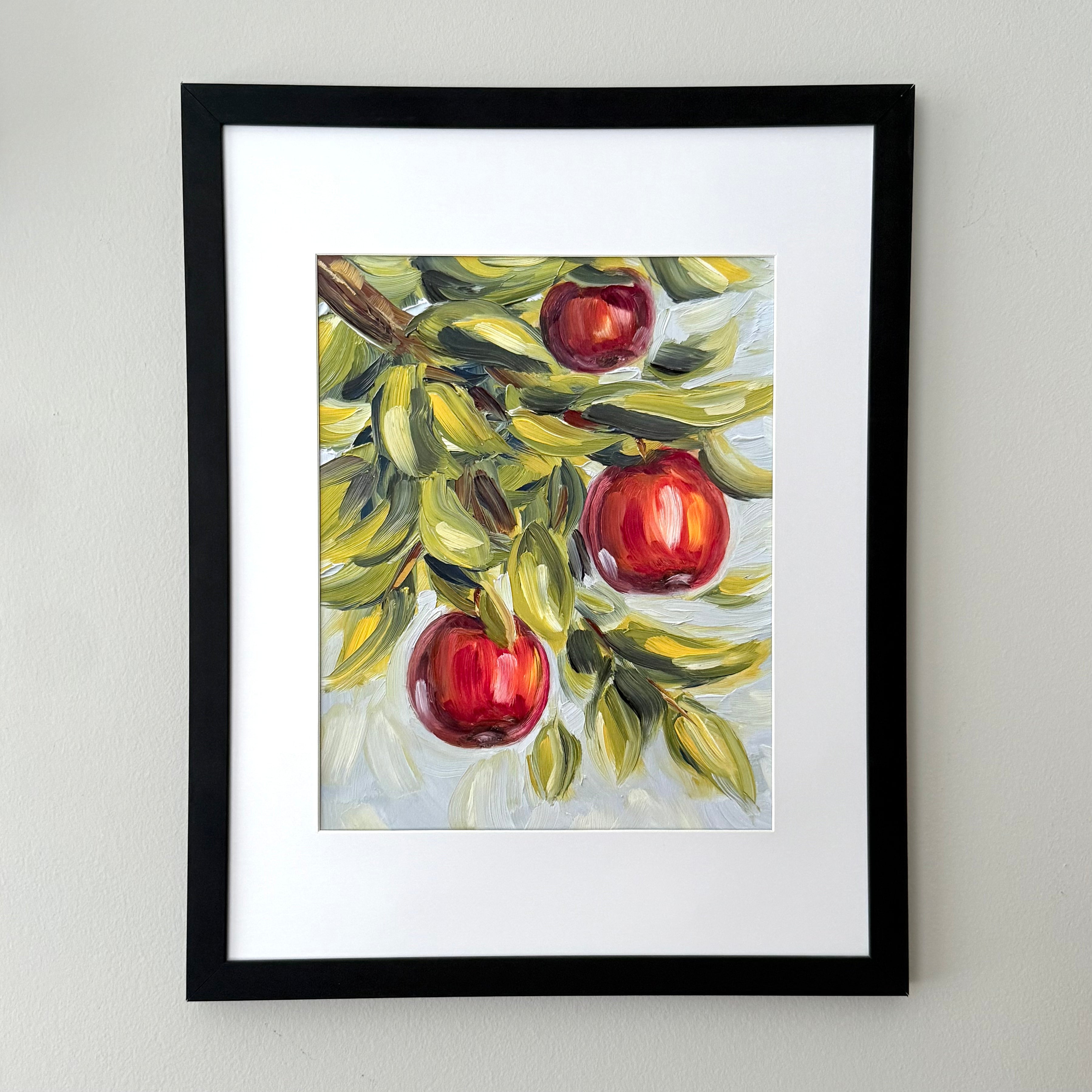 A framed art print featuring a painted still life of three red apples on a branch with green and yellow leaves, set against a light gray wall.