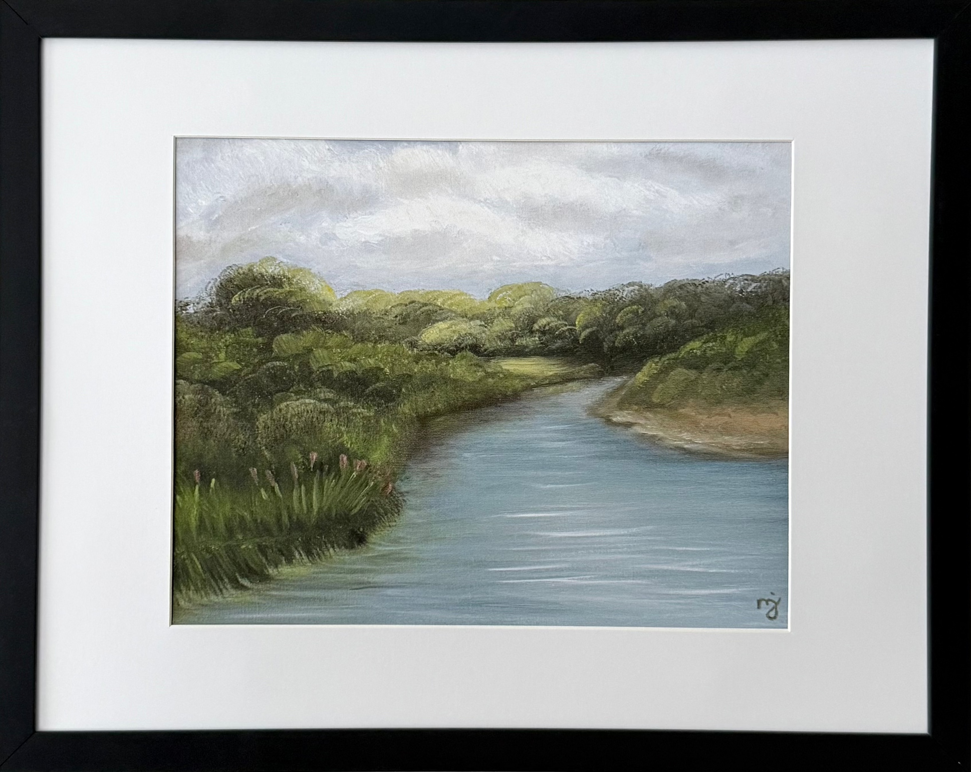 A framed landscape art print depicting a winding river bordered by green trees and grass under a cloudy sky, mounted on a light beige wall.