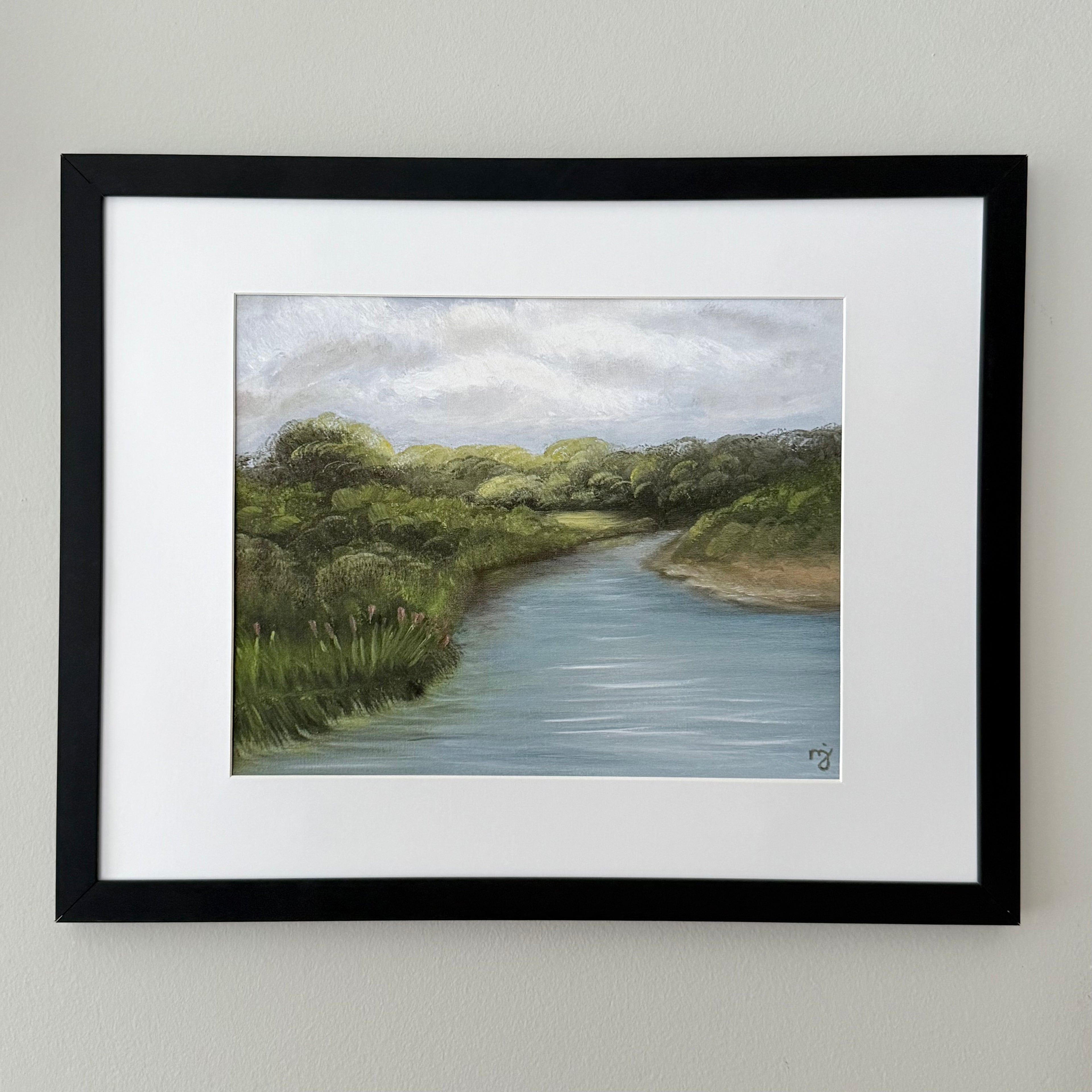 A framed landscape art print depicting a winding river bordered by green trees and grass under a cloudy sky, mounted on a light beige wall.