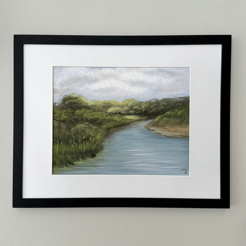 A framed landscape art print depicting a winding river bordered by green trees and grass under a cloudy sky, mounted on a light beige wall.