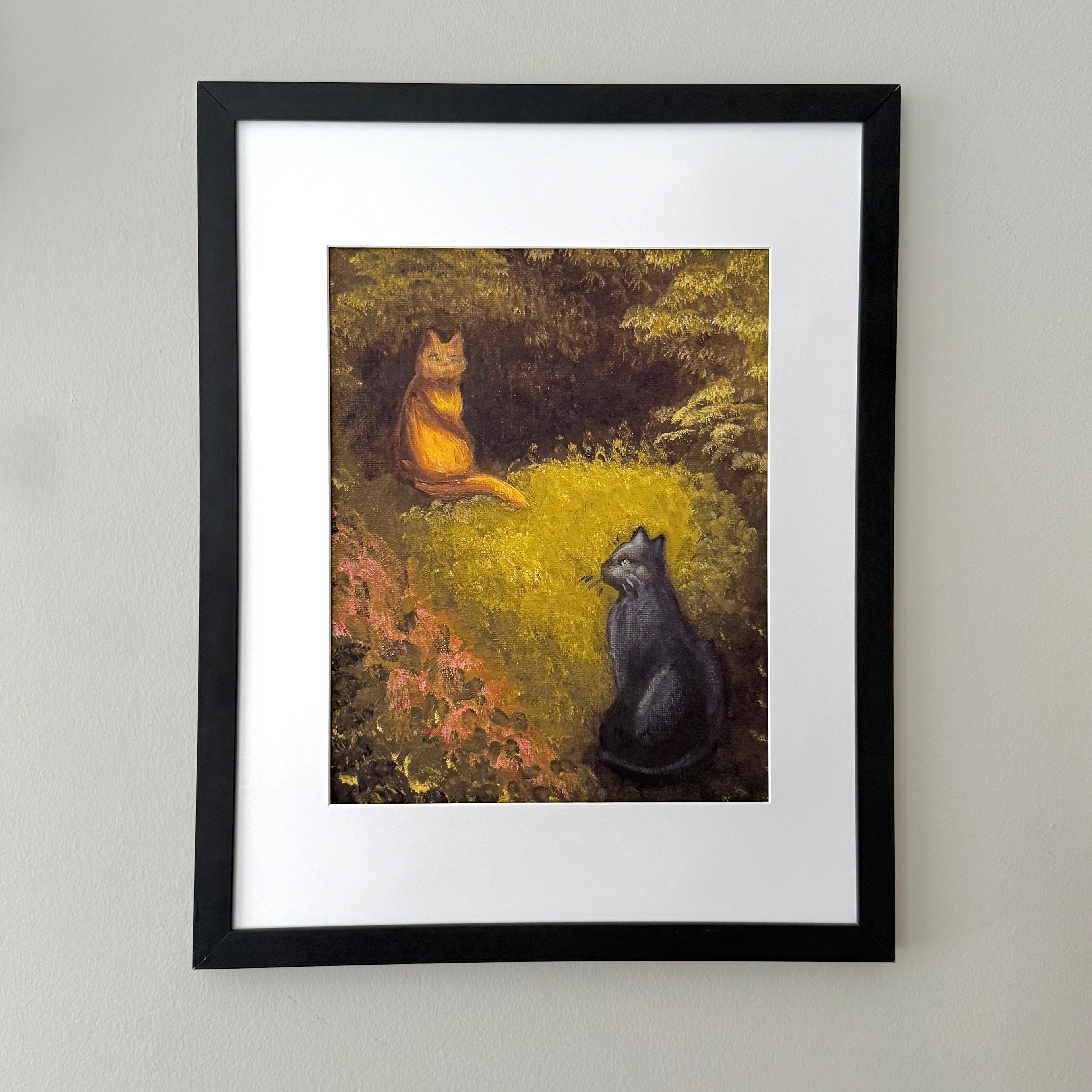 Framed art print featuring a painting of two cats, one orange and one black, sitting in a grassy field, displayed on a light gray wall.