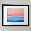 Framed landscape art print featuring layered blue mountains under a pink and orange sky, mounted on a white mat within a black frame against a light gray wall.