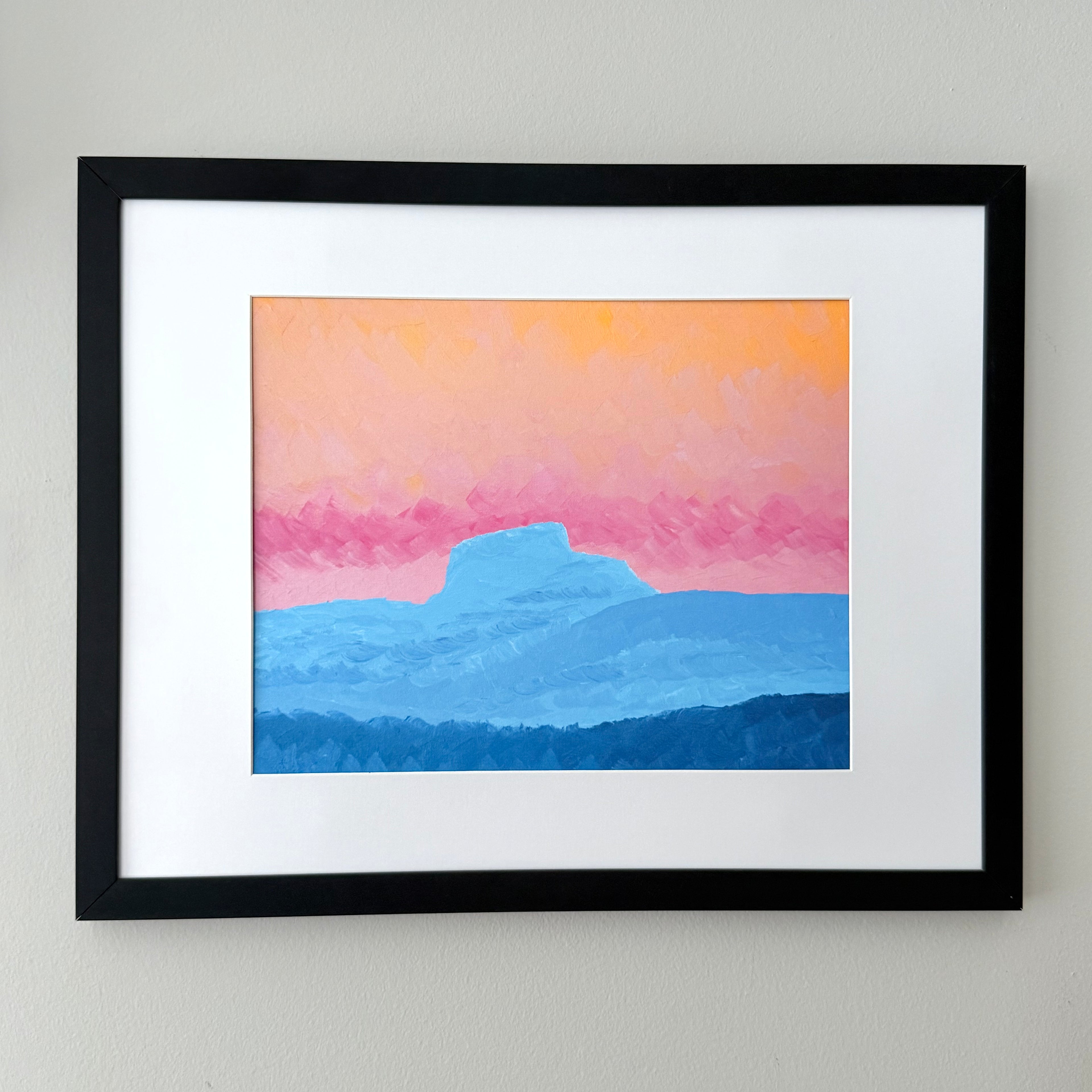 Framed landscape art print featuring layered blue mountains under a pink and orange sky, mounted on a white mat within a black frame against a light gray wall.