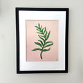 Framed botanical art print featuring a green leaf illustration on a soft pink background, matted with white and enclosed in a black frame, mounted on a light gray wall.