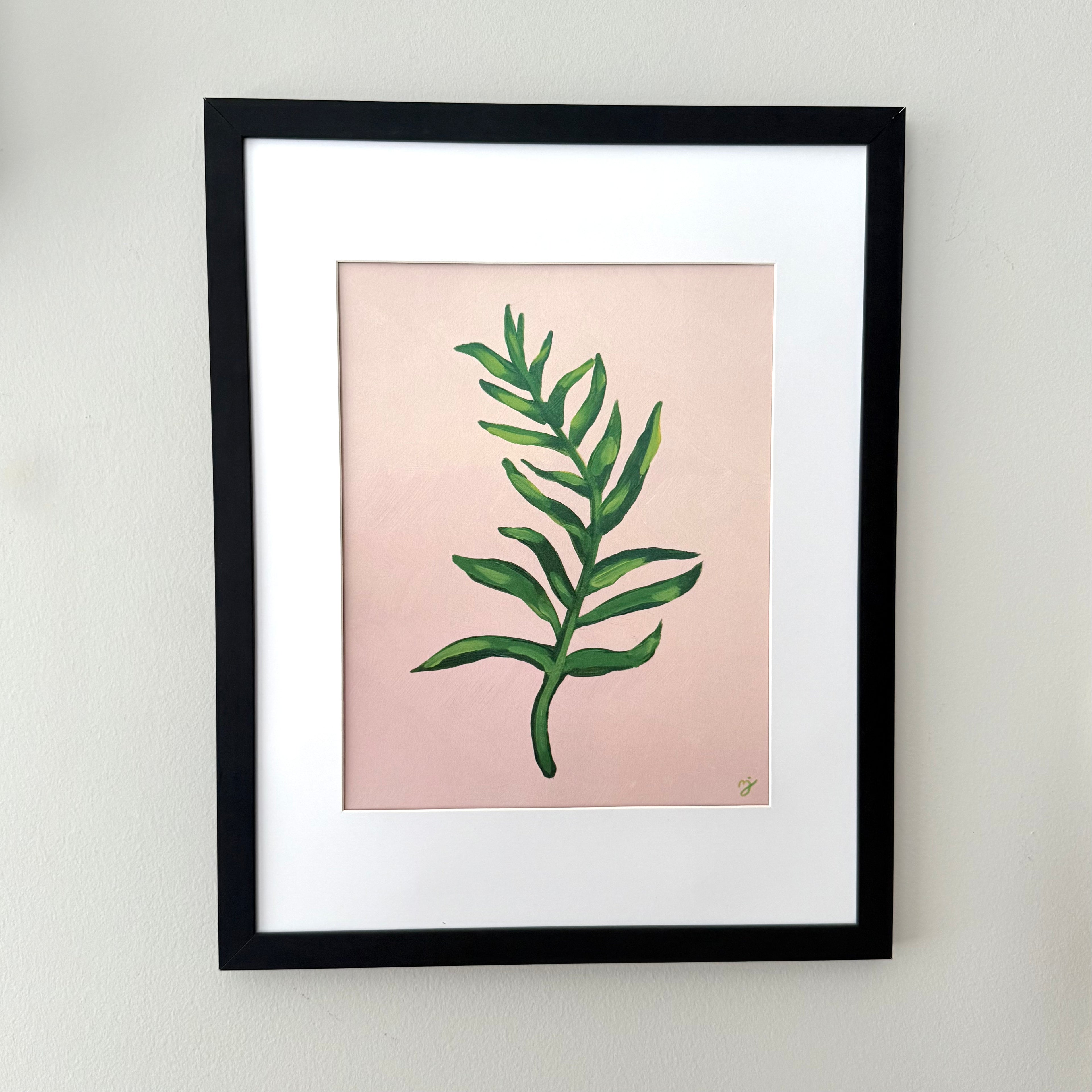 Framed botanical art print featuring a green leaf illustration on a soft pink background, matted with white and enclosed in a black frame, mounted on a light gray wall.