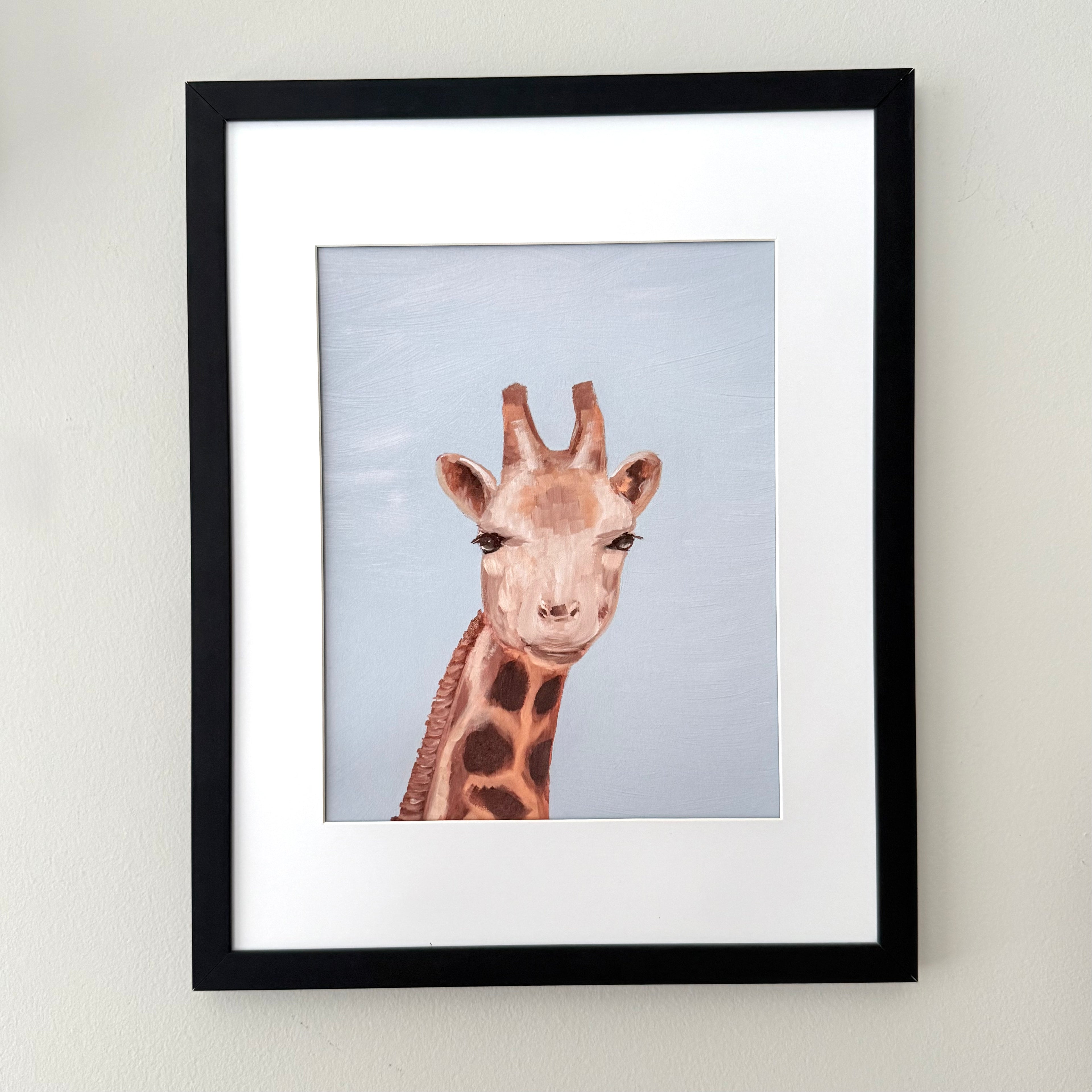 Framed wall art featuring a painted giraffe portrait with a light blue background, set in a black frame with a white mat against a neutral wall.