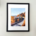 A framed landscape painting of a winding road through autumn trees, displayed on a light beige wall. The artwork features a light blue sky, vibrant orange and yellow foliage, and dark green pine trees, enclosed in a white mat and a simple black frame.