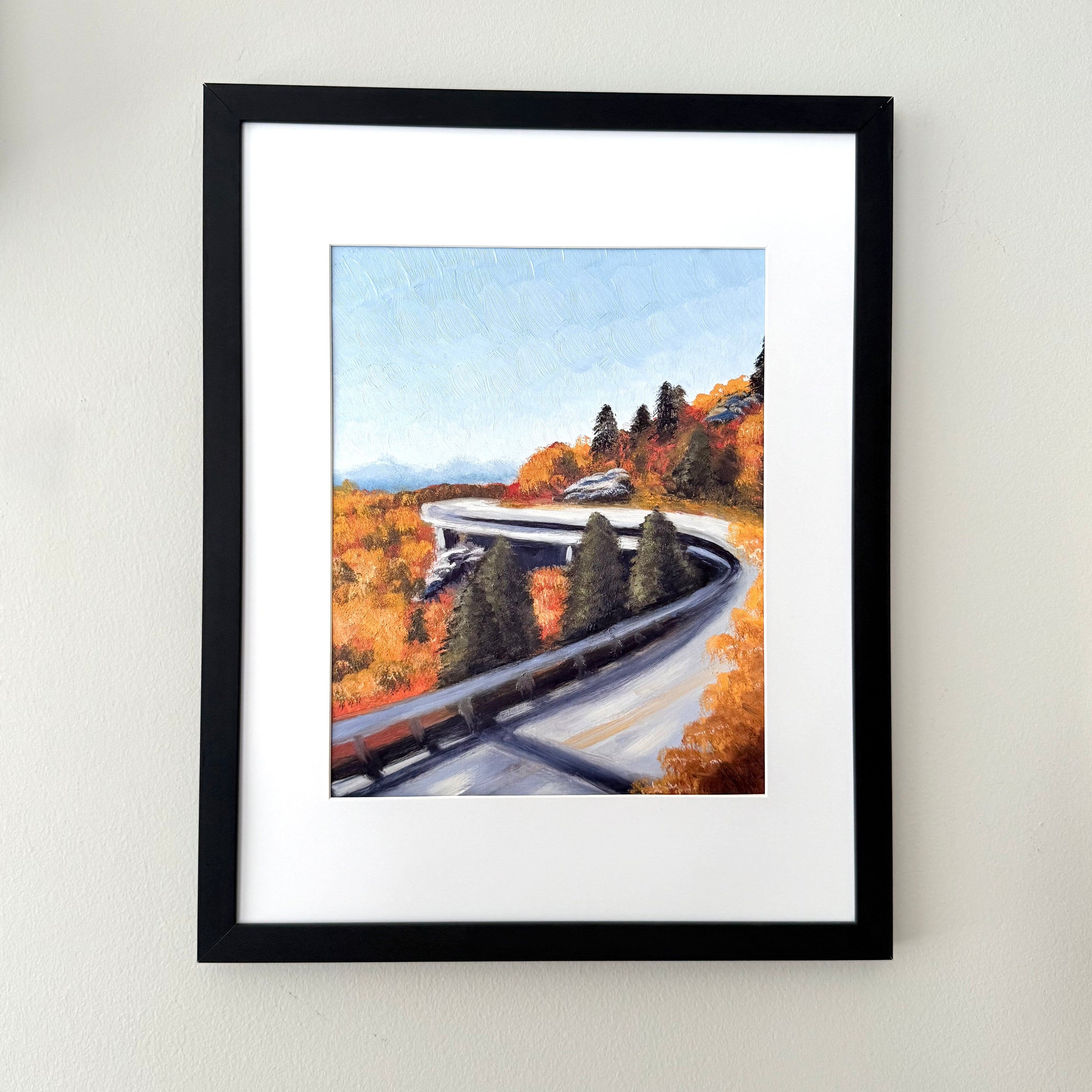 A framed landscape painting of a winding road through autumn trees, displayed on a light beige wall. The artwork features a light blue sky, vibrant orange and yellow foliage, and dark green pine trees, enclosed in a white mat and a simple black frame.