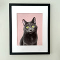 Framed art print featuring a portrait of a black cat with bright yellow-green eyes on a dusty pink background, matted in white and housed in a black frame, mounted on a light gray wall.