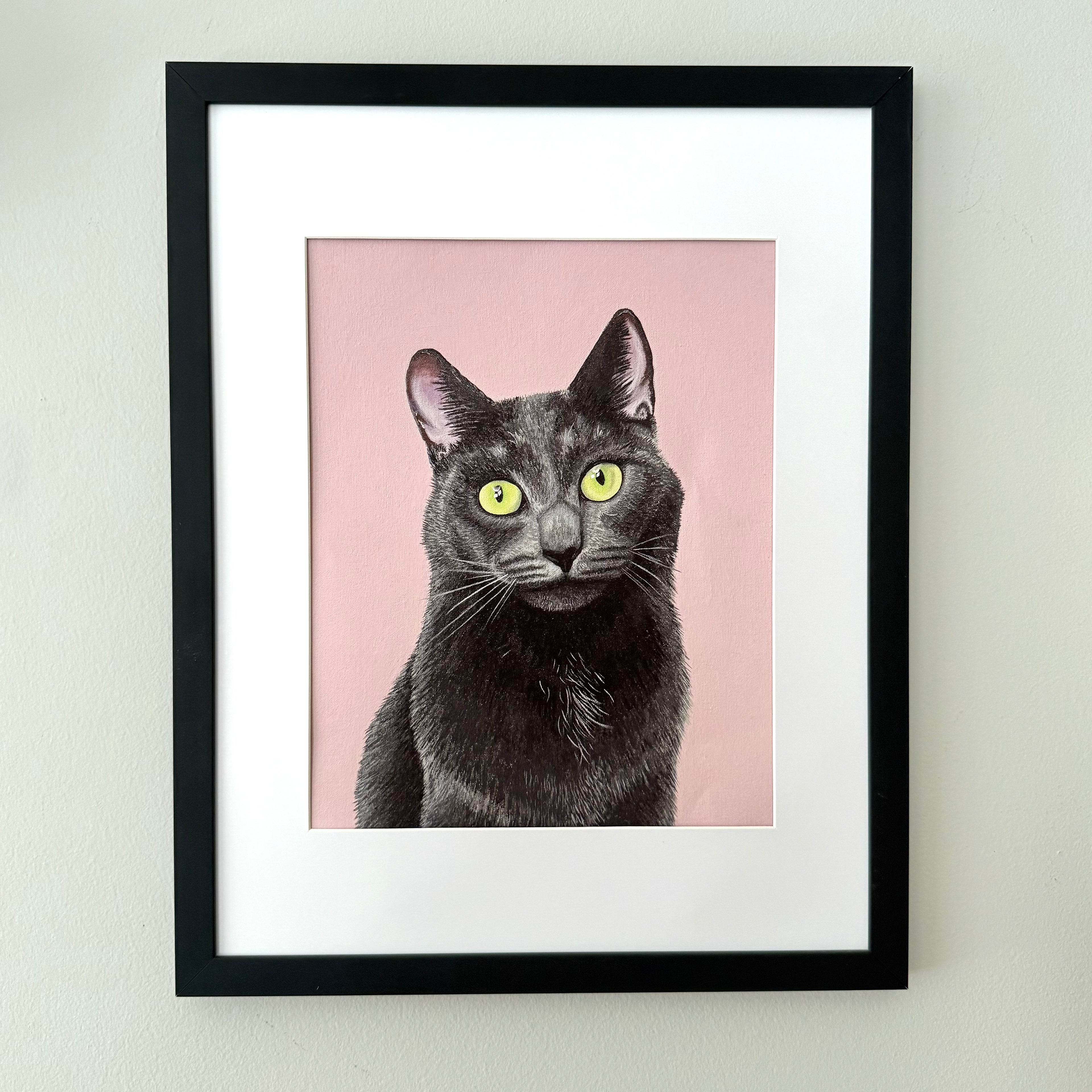 Framed art print featuring a portrait of a black cat with bright yellow-green eyes on a dusty pink background, matted in white and housed in a black frame, mounted on a light gray wall.