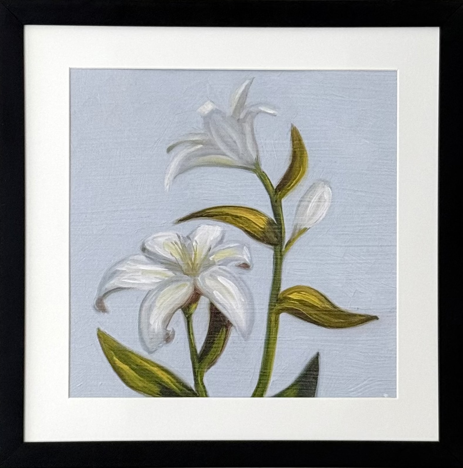 A framed botanical art print of white lilies with green leaves on a light blue-gray background, presented in a black frame with a white mat against a neutral beige wall.