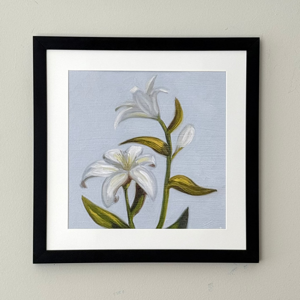 A framed botanical art print of white lilies with green leaves on a light blue-gray background, presented in a black frame with a white mat against a neutral beige wall.
