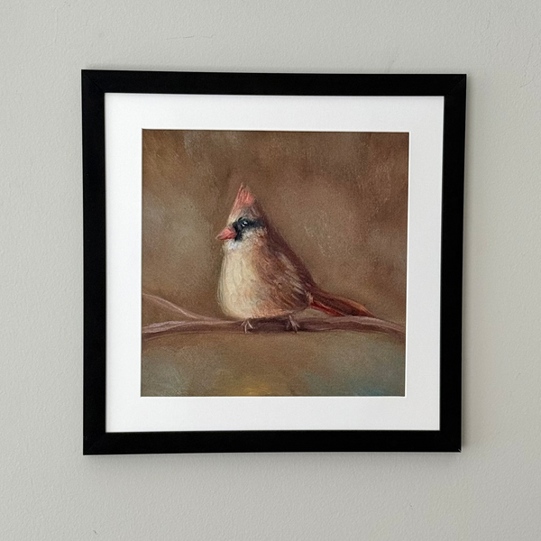 A framed art print featuring a painted cardinal perched on a branch, displayed on a light gray wall. The artwork has a brown and beige background with the cardinal in brown, beige, and red tones, surrounded by a white mat and a black frame.